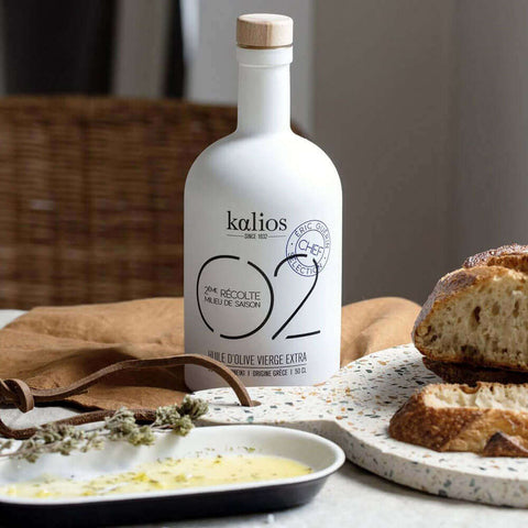 Bottle of Extra Virgin Olive Oil 02 of the Kalios brand, placed on a table, with a wooden board containing bread and sauce, front view. 