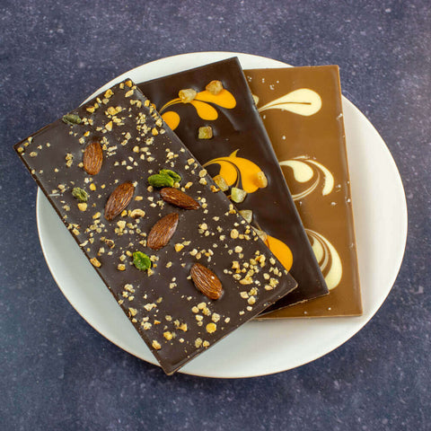 A Dark Chocolate With Dried Fruits Bar, a Dark Chocolate Marbled With Orange Peel Bar and a Milk Chocolate With White Caramel Marbled With Salt Flower Bar, all from the Ile De R?? Chocolats brand, arranged on a plate, seen from above.