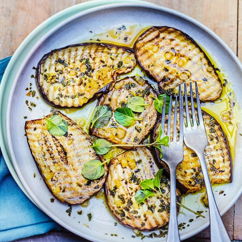 Assorted cooked and sliced eggplant, arranged on a round plate with some herbs, seen from above. 