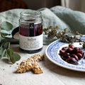 Glass jar containing Greek Kalamata Olives Natural of the Kalios brand opened with some olives in a plate and with breadstick next to it, front view. 