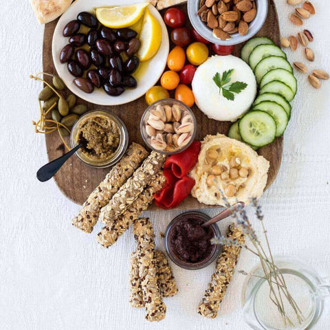 Wooden tray containing olives, terrine, tomatoes, cucumbers, fines, and 7 Cereals Breadsticks, seen from above. 