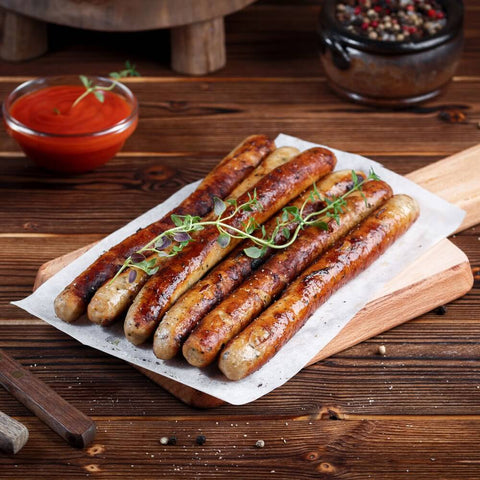 Chipolata Bistro Sausage cooked and placed on a sheet of baking paper on a wooden board, with a jar of sauce next to it, seen from the front.
