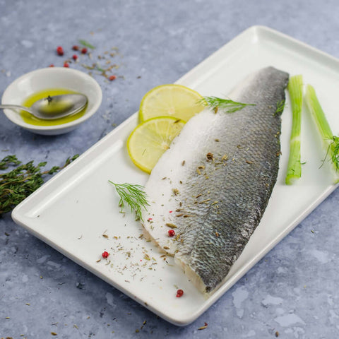 Piece of Estuary Branzino - European Seabass (Skin-On) in a rectangle dish with lemon, chives, and pink peppercorns, seen from above.