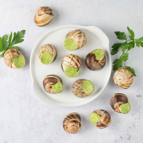 Assortment of 12 Escargots in Parsley-Garlic Butter, 6 of them on a round plate, the rest on a marble table, with some basil leaves, seen from above. 