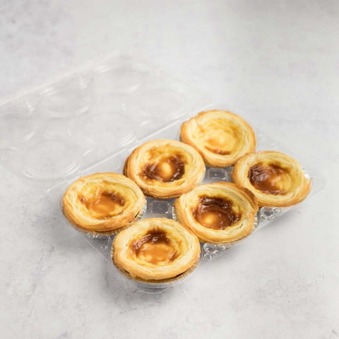 Assortment of 6 Pastéis de Nata arranged in their plastic tray, placed on marble, seen from above.