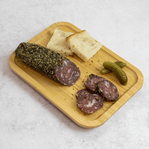 A wooden board with two pieces of bread, two pickles and sliced French Pork Salami With Herb, seen from above.