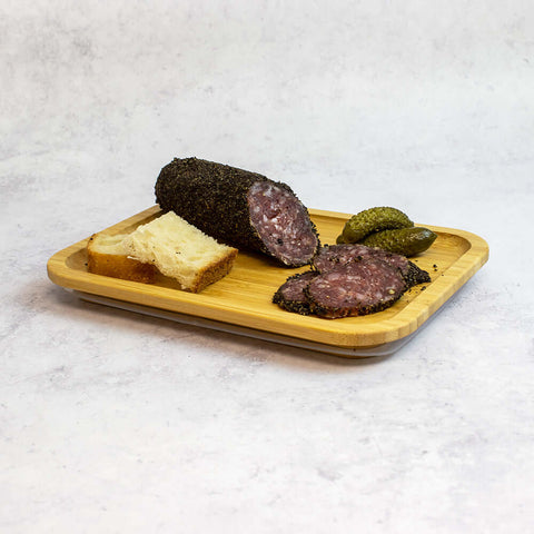 A wooden board with two pieces of bread, two pickles and sliced French Pork Salami With Black Pepper, seen from the front.