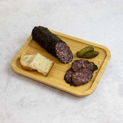A wooden board with two pieces of bread, two pickles and sliced French Pork Salami With Black Pepper, seen from above.