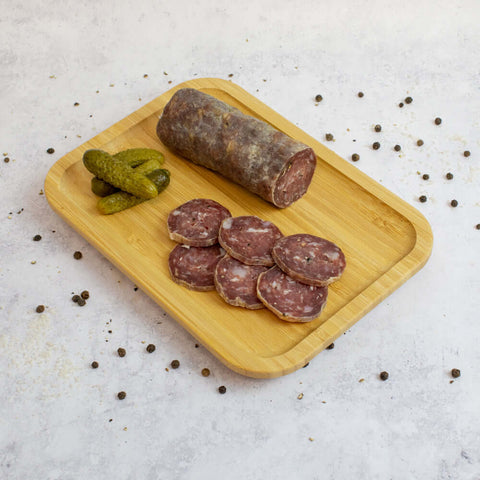 A wooden board with three pickles and sliced French Pork Salami with some black peppercorns next to it, seen from above.