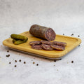 A wooden board with three pickles and sliced French Pork Salami with some black peppercorns next to it, seen from the front.